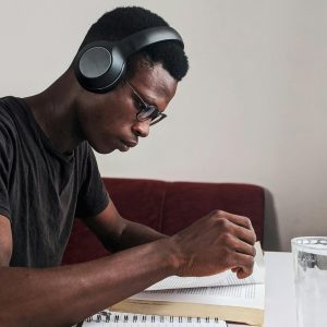 An adult man intensely studying with headphones, pen, and notebook at a desk indoors.