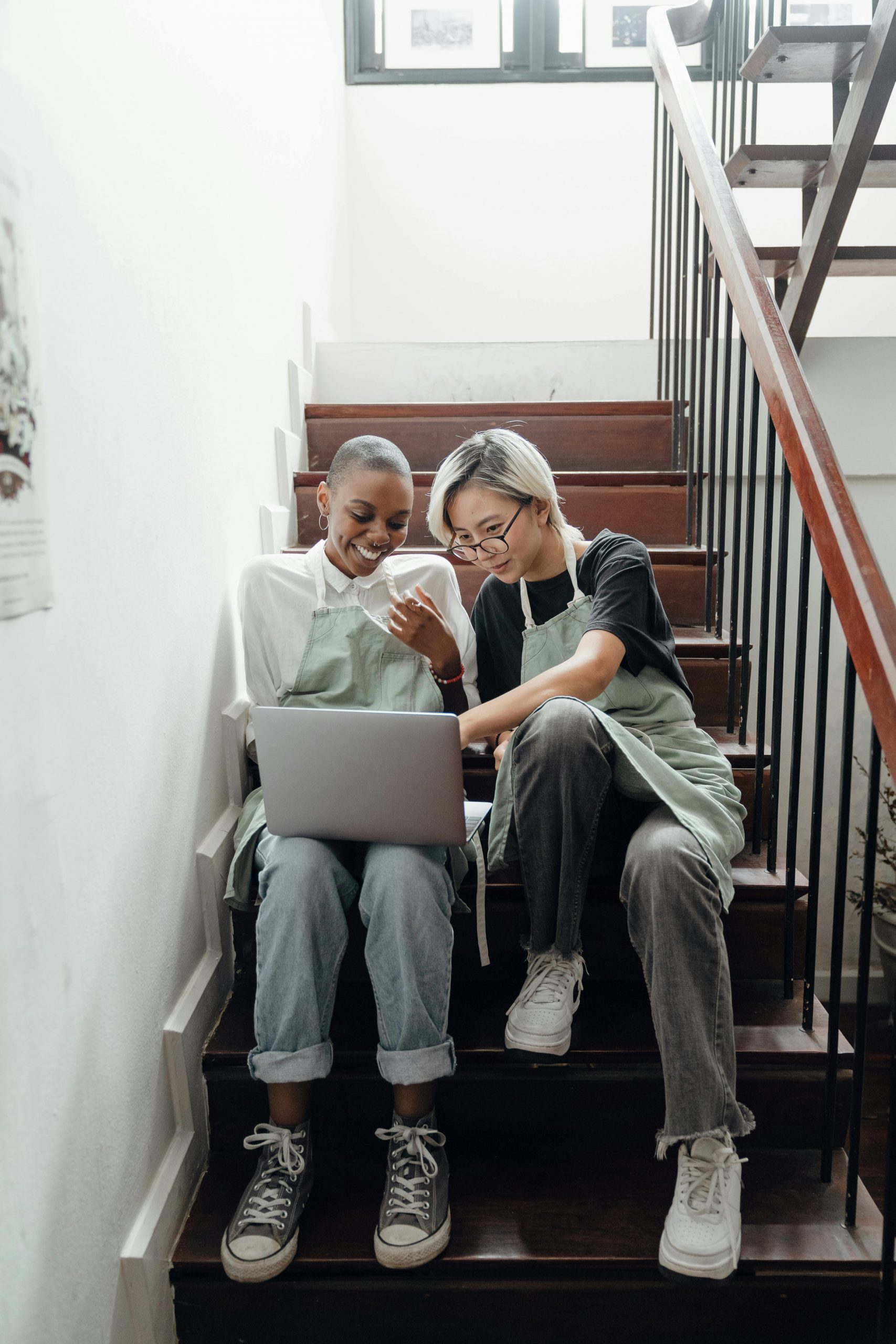 Full body of positive young diverse female friends in trendy clothes and aprons watching funny video on laptop sitting on staircase