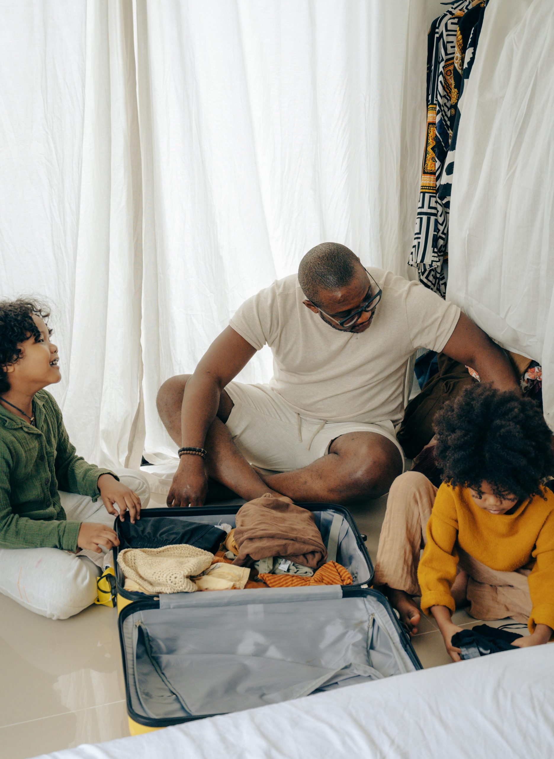 From above of young African American man sitting on floor and packing suitcase with little daughters