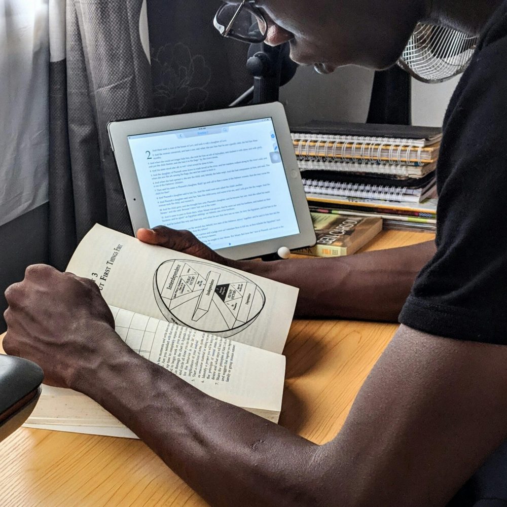 A man studying with a book and tablet at a desk indoors, showcasing multitasking and modern study habits.