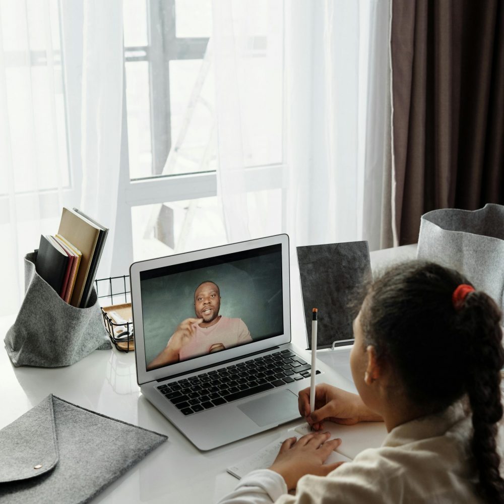 A young girl takes notes during an online class while using a laptop at her study desk.