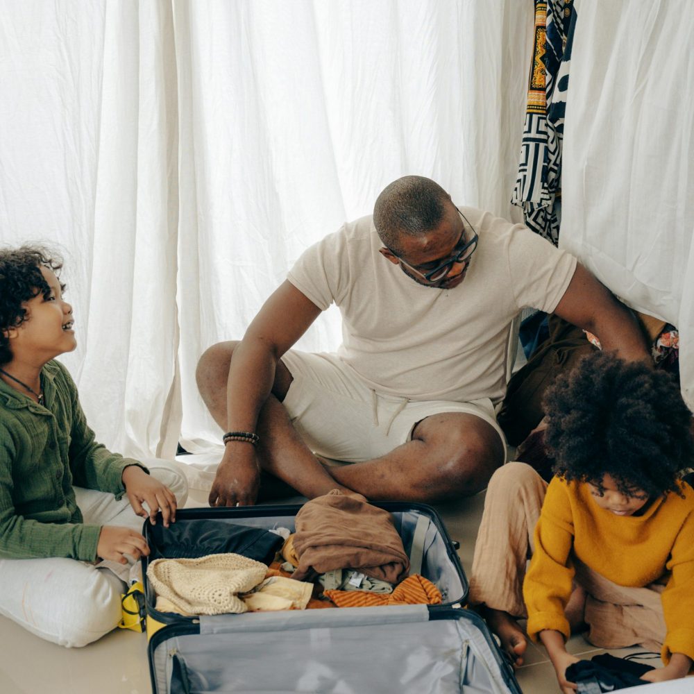 From above of young African American man sitting on floor and packing suitcase with little daughters