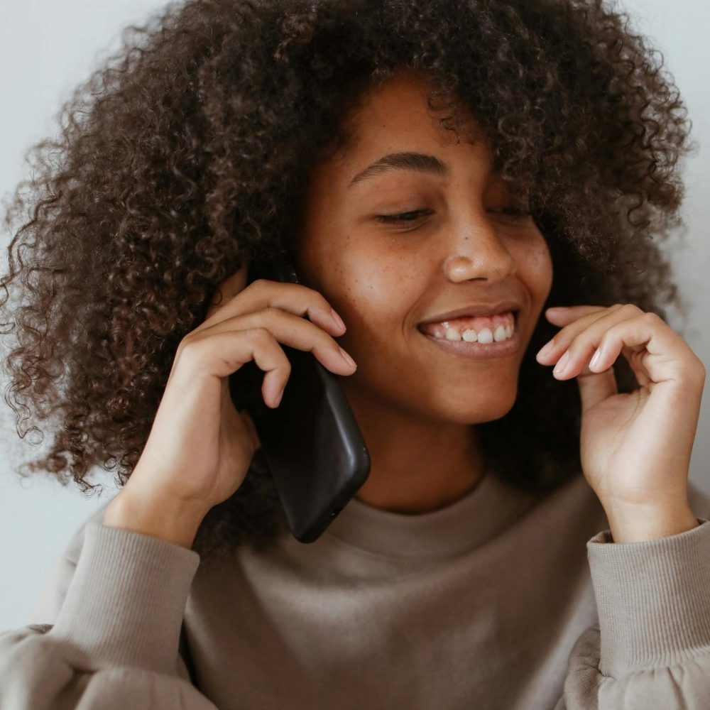 Smiling woman with afro hair enjoying a phone call indoors, wearing a casual sweater.