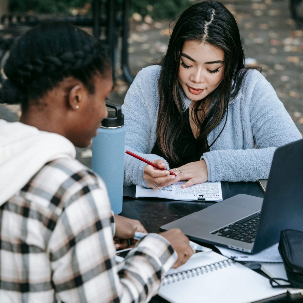Two diverse students collaborating on a project outdoors with laptops and notes on a college campus.